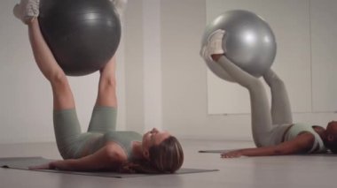 Sırtüstü uzanan ve bacaklarıyla topları kaldıran iki genç ve güçlü kadın kapalı stüdyoda spor yapıyorlar.