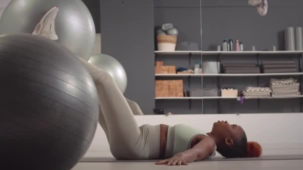 在工作室的室内锻炼中 身穿紧身衣的强壮的年轻黑人妇女靠着背躺着 双腿提起健身球 — 图库视频影像