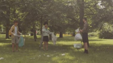 Erkek öğretmenin çeşitli okul çocuklarıyla birlikte güneşli bir günde halka açık parktaki çimlerde çöp toplarken tam boy görüntüsü.