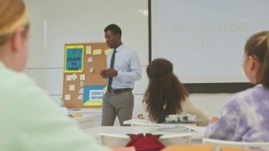 Hevesli genç siyahi erkek öğretmen çeşitli 6. sınıf öğrencilerine etkileşimli bilişim dersi veriyor. Parlak ferah sınıflarında masalarında oturuyorlar ve kod yazmayı öğreniyorlar.