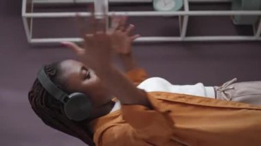 Genç Afrikalı Amerikalı bir kadının evde tek başına otururken kulaklıkla müzik eşliğinde dans edişinin dikey görüntüsü.