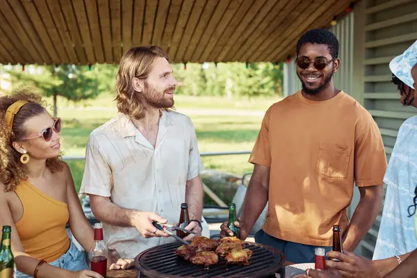 Portrait of two young men grilling meat outdoors with diverse group of friends at barbeque party in Summer
