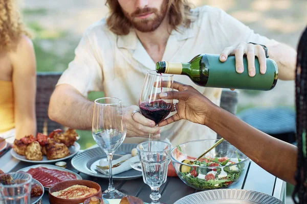Close up of couple pouring red wine into glass while enjoying dinner party with friends outdoors in Summer