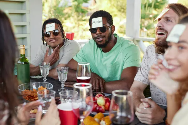 Portrait of black man with sticker note on head playing Guess who game with diverse group of friends at outdoor party