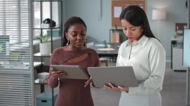 Bilgisayarlı ve dijital tabletli iki genç bayan meslektaşın ofiste sohbet ederken orta boy görüntüsü.
