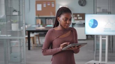 Elinde dijital tabletle ofiste kameraya poz veren Afro-Amerikalı genç iş kadınının orta boy portresi.
