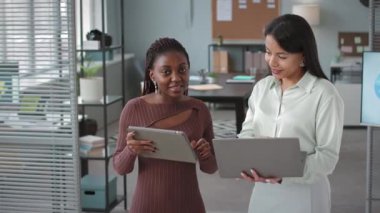 Modern ofiste kameraya poz veren iki genç, etnik çeşitliliğe sahip bayan meslektaşın orta boy portresi.