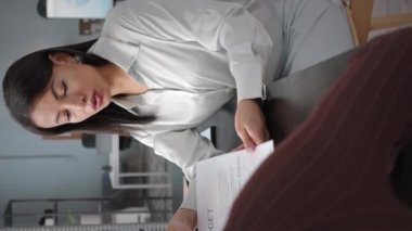 Genç, esmer, çift ırklı bir kadının modern ofisteki kadın meslektaşıyla şirket bütçesi ve finansmanı hakkında konuştuğu dikey bel resmi.