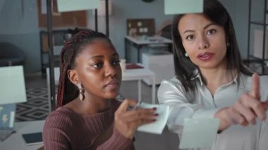 İki genç, yaratıcı, etnik çeşitlilikte kadın meslektaşın fikir alışverişinde bulunup iş planlarını tartışırken cam pencereye etiket yapıştırması.