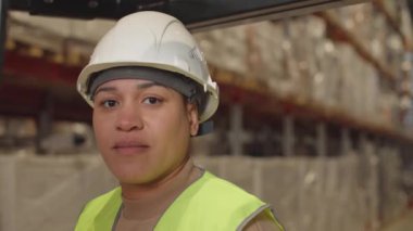 Çift ırklı kadın forklift sürücüsünün beyaz şapkalı orta boy portresi. Büyük depoda çalışan kameraya bakıyor. Paletlerde karton kutular ve bulanık arka planda raflar var.