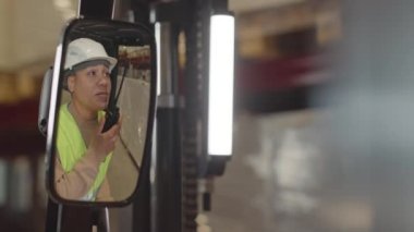 Dikiz aynasından, güvenlik kaskı ve asit yeşili yelek giyen genç çift ırklı kadın operatörün depoda birlikte çalışırken çalışanlarına talimat verirken çekilmiş.