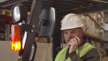 Depoda iş arkadaşlarıyla konuşurken kameraya bakan olgun beyaz forklift operatörünün göğüs portresi.