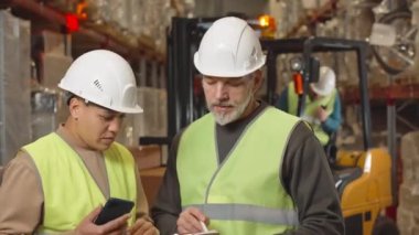 Çok ırklı iki kadın ve erkek depo çalışanının orta boy portresi forklifte karşı duruyor. Uyarı ışığı ve operatör sürücü koltuğundan iletici ile talimatlar veriyor.