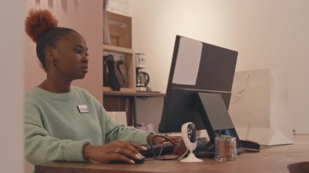 若い黒人女性がオンライン販売をチェックするカウンターでコンピュータが立ち並ぶ高級デザイナー服店のショップアシスタントとして働くのを待つ — ストック動画