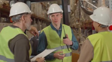 Beyaz tenli erkek forklift operatörü ve iki iş arkadaşının orta boy fotoğrafları. Hepsi de depoda nöbet tutarken başlık ve asit yeşili yeleklerle sohbet ediyorlar.
