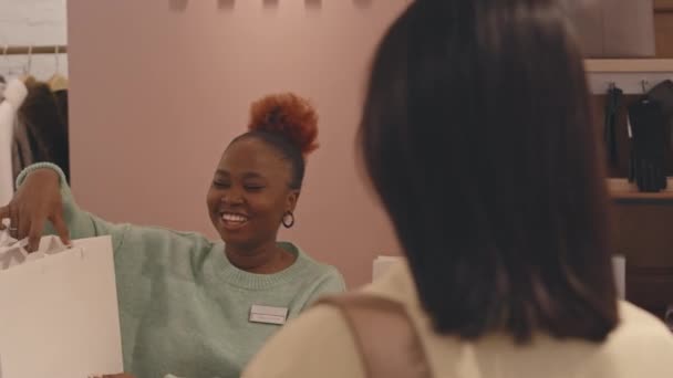 积极的非洲裔美国女售货员将白纸购物袋送给身穿名牌服装精品店购物的年轻优雅的白人妇女 — 图库视频影像