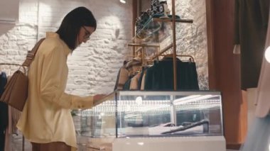 Lüks giyim mağazasından alışveriş yaparken tasarım mücevherleri ve aksesuarlarla LED aydınlatma vitrinini inceleyen zarif beyaz bir kadın.