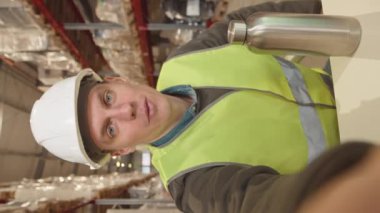 Kameradaki konuşma sırasında genç beyaz erkek depo işçisinin sabit şapka ve asit yeşili yelek giyip video blogu kaydettiği dikey el kamerası görüntüsü.