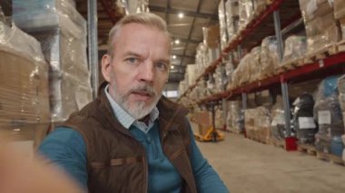 Yetişkin beyaz erkek depo yöneticisinin el bilgisayarı kamerası önünde konuşurken iş yerinde video blogu kaydediyor.