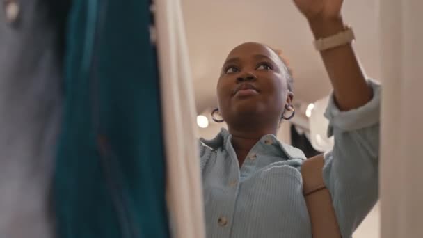 高級ブティックで買い物しながらラックのスタイリッシュな服を通して見る陽気な若いアフリカ系アメリカ人女性の低い角度 — ストック動画