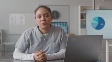 Çağdaş tıp ofisinde masasında oturan ve kameraya konuşan önlüklü genç çift ırklı kadın hemşirenin bel ponksiyonu.