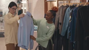 Orta boy Afro-Amerikalı bayan tezgahtar tasarımcı dükkanında süveter seçen genç bir kadına danışıyor.