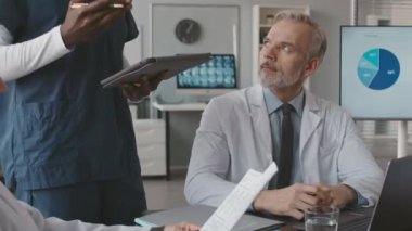 Tanımlanamayan, dijital tabletli siyahi erkek hemşirenin resmi. Ofiste toplantı yaparken yetişkin beyaz erkek doktorla görüşüyor.