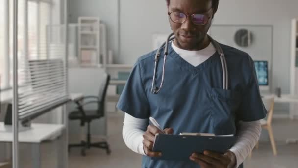 将年轻快乐的黑人男性医生的画像倾斜起来 他肩上戴着蓝色的刷子和听诊器 在剪贴板上做笔记 并摆出姿势 站在明亮的现代医生办公室里拍照 — 图库视频影像