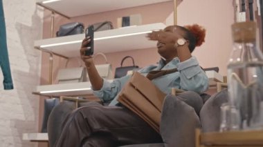 Modern tasarımcı butiğinde rahat bir bölgede otururken akıllı telefondaki genç siyahi kadın görüntülerinin orta boy görüntüsü.