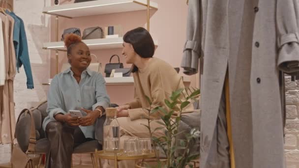 ショッピング中に会話をするデザイナーブティックで快適なアームチェアに座っている2人の若い幸せな多民族女性の親友 — ストック動画