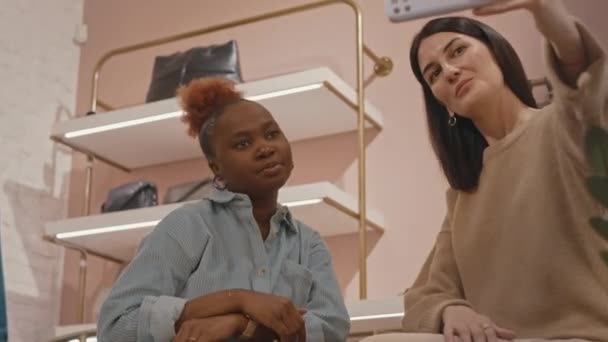デザイナー服店で買い物中にスマートフォンでセルフィーの肖像画を撮る2人の若い多様な女性の親友の低い角度 — ストック動画