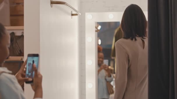 女性の友人がスマートフォンでビデオを録画しながら 現代の服店のフィッティングルームに鏡の前にポーズするエレガントなドレスの若い美しい白人女性のミディアムスローモ — ストック動画
