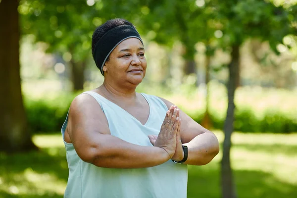 Waist up portrait of mature black woman doing yoga outdoors in green park and looking away with mindfulness, copy space