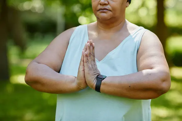 Cropped portrait of mature black woman doing yoga outdoors in green park and holding hands together for balance in prayer position, copy space