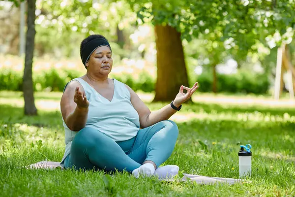 Full length portrait of overweight black woman doing yoga outdoors and meditating with eyes closed on green grass