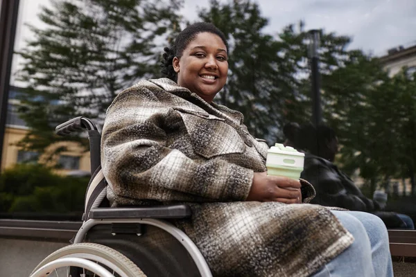 Portrait of young black woman in wheelchair smiling at camera in city setting and holding coffee cup