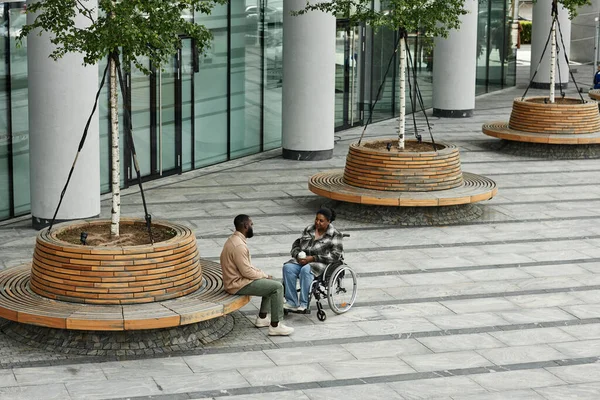 Wide angle view at adult black couple with partner in wheelchair chatting outdoors in city setting, graphic tiled floor, copy space
