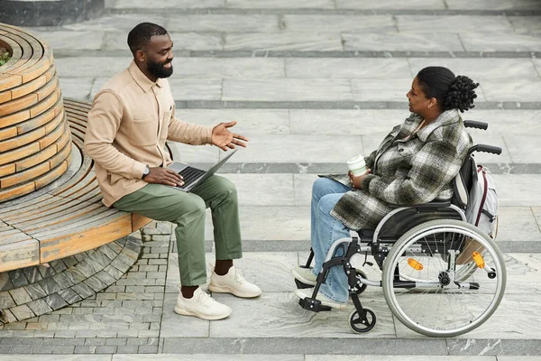Full length shot of adult black couple with woman in wheelchair chatting outdoors in city setting and enjoying coffee