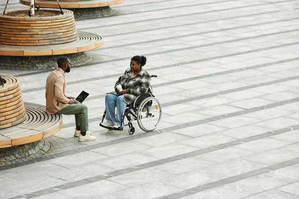 Graphic wide angle view of adult couple with black woman in wheelchair chatting outdoors in city setting against tiled floor, copy space