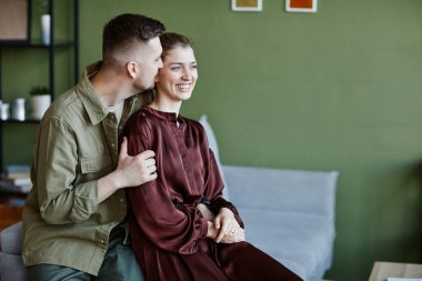 Genç adam kız arkadaşını kucaklıyor ve öpüyor onlar oturma odasında kanepede otururken