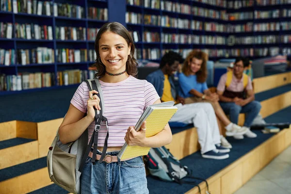 Waist up portrait of young woman with backpack standing in library and looking at camera, copy space