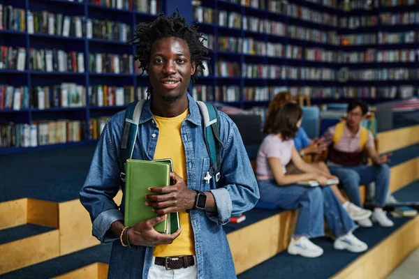 Vibrant waist up portrait of black young man holding books in college library and looking at camera, copy space