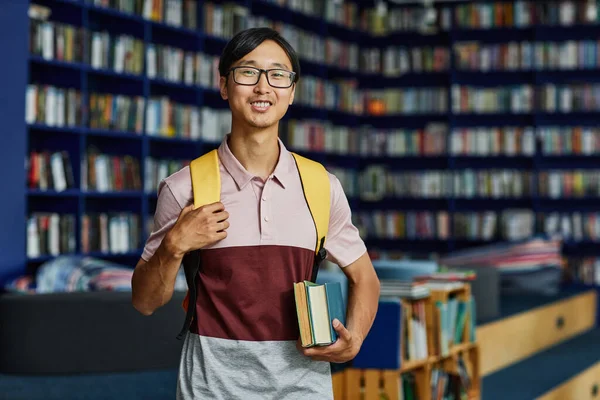 举着背包站在图书馆里的亚洲年轻人的肖像 微笑着看着相机 复制空间 — 图库照片