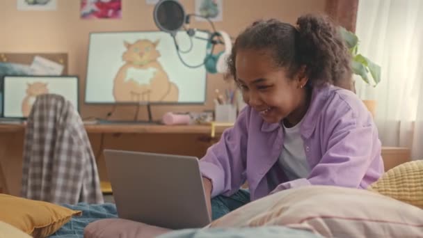 穿着时髦薰衣草衬衫的快乐的非洲裔美国女孩坐在柔软的床上 躺在明亮多彩的卧室里 与朋友们在笔记本电脑上发短信 — 图库视频影像