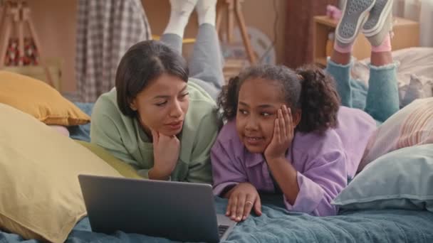 アフリカ系アメリカ人の母親と遺伝子Zの娘は 明るい子供の寝室で柔らかいベッドで胃の上に横たわり ラップトップでチャットやブラウジング — ストック動画