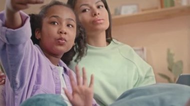 Neşeli Afro-Amerikalı, çocukluk çağında, annesi evde otururken akıllı telefondan fotoğraf çektirirken komik suratlar yapan bir kız.