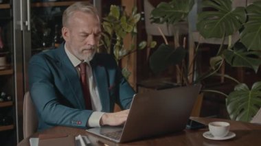 Kafkas sakallı, takım elbiseli bir iş adamının restoran masasında oturmuş dizüstü bilgisayarının klavyesinde otururken belini kaldırması.