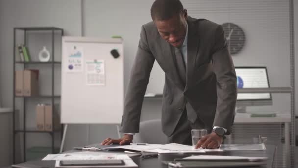 オフィスデスクに立ってカメラを見ながらビジネス文書を分析する正式なスーツで若いアフリカ系アメリカ人のビジネスマンの中型肖像画 — ストック動画