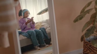 Afro-Amerikan ergen bir kızın, kablosuz kulaklıklı, akıllı telefonuyla yatak odasında boş vakitlerinde otururken çekilmiş uzun bir ayna görüntüsüyle.