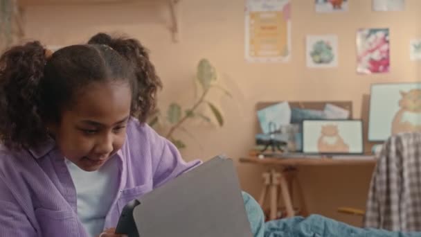 かわいいアフリカ系アメリカ人 歳の少女は カラフルなキッズベッドルームでベッドに座っている間 デジタルタブレットでゲームをプレイ — ストック動画